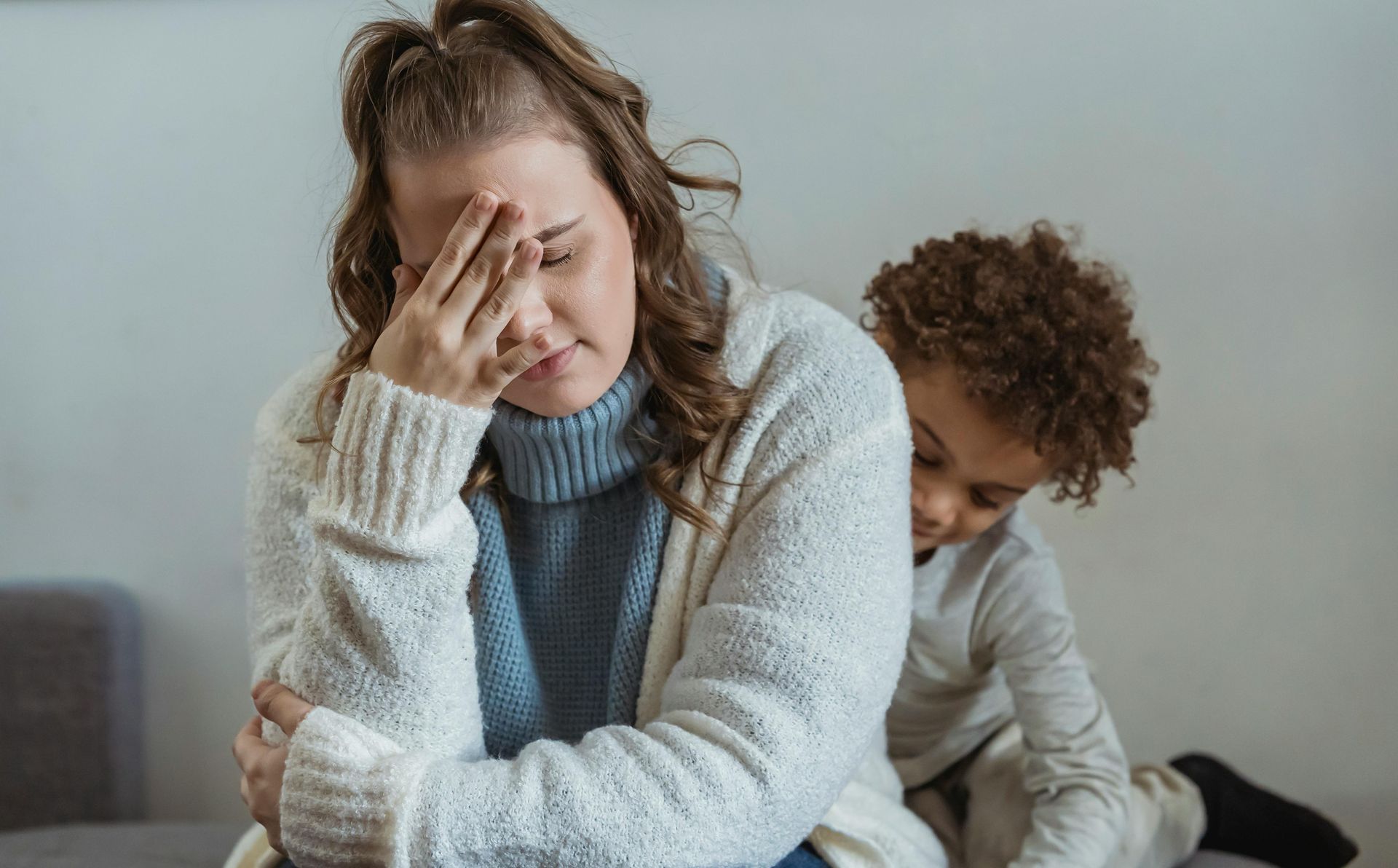 Woman with hand on forehead looks stressed; child clings to her back.
