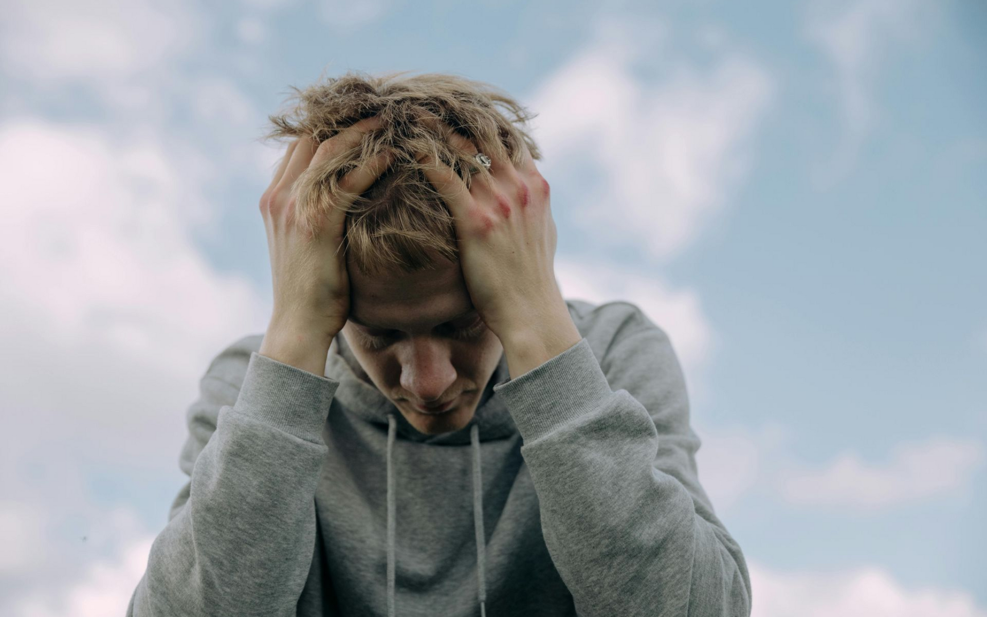 Person with light hair, hands on head, looking down, set against a cloudy blue sky.