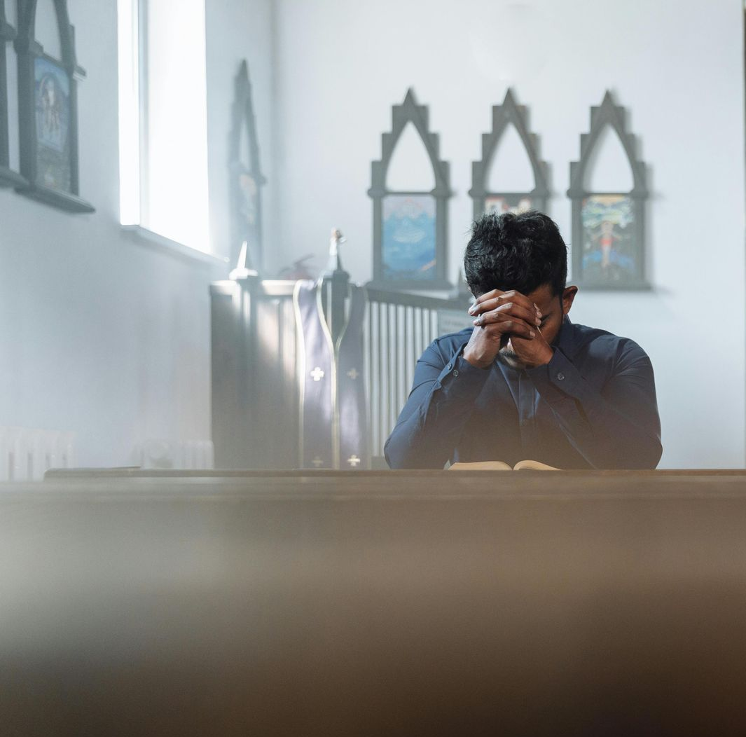Man praying at a church, hands clasped. He is sitting at a wooden pew, next to a window.