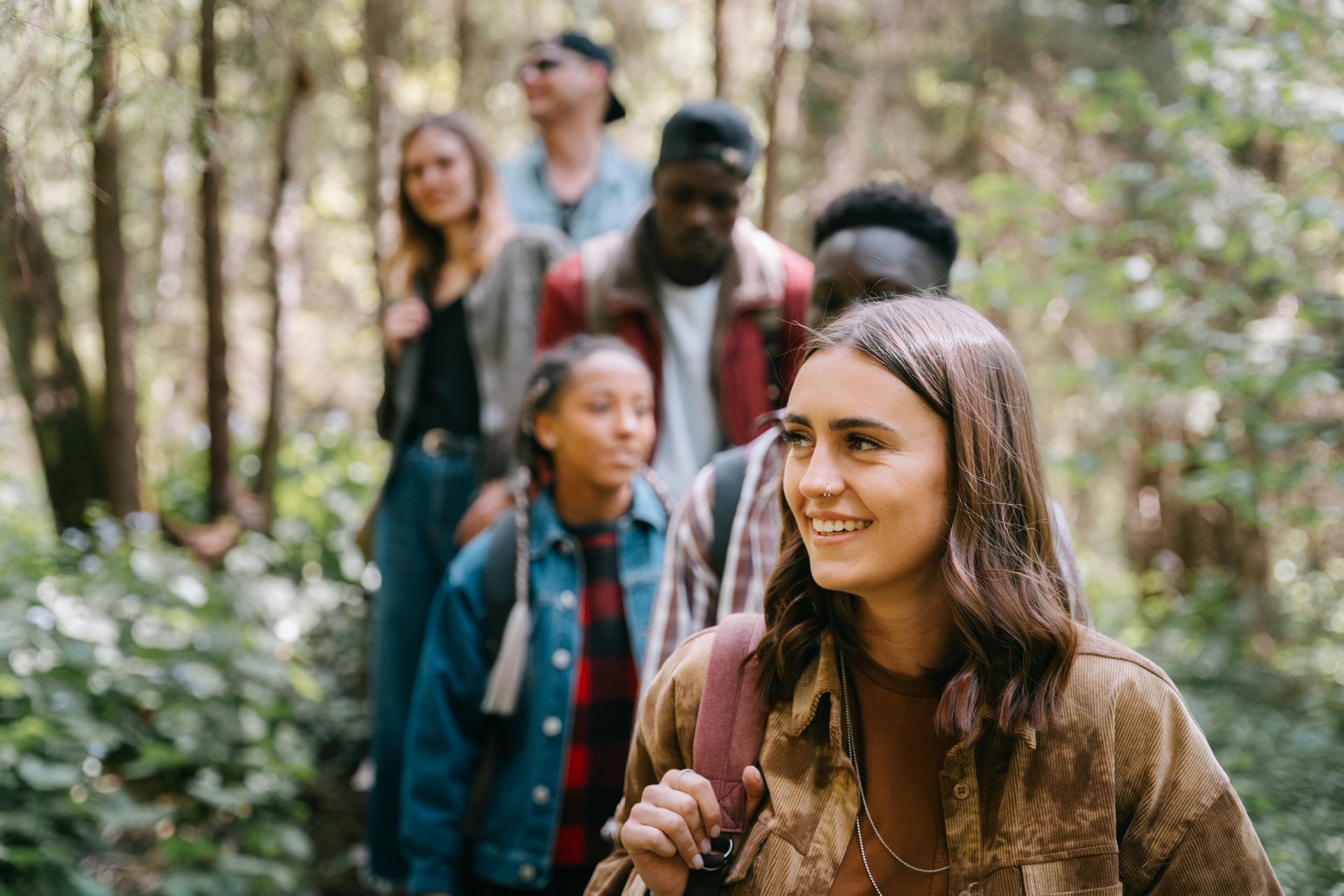 Group of people hiking through a forest; smiling woman in front, others follow.
