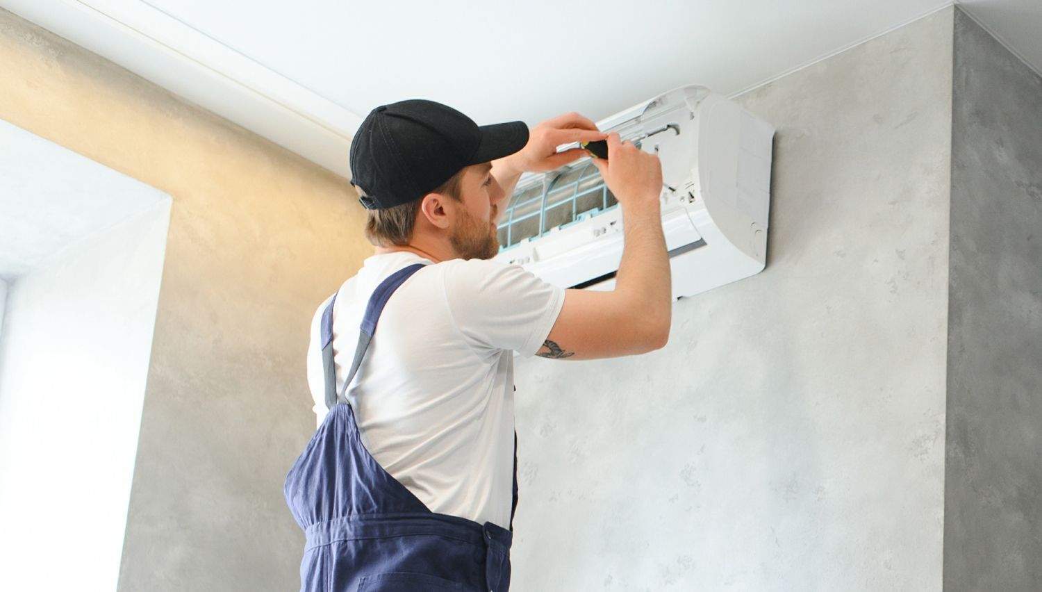 A man in overalls and a cap repairs a wall-mounted air conditioner with a screwdriver.