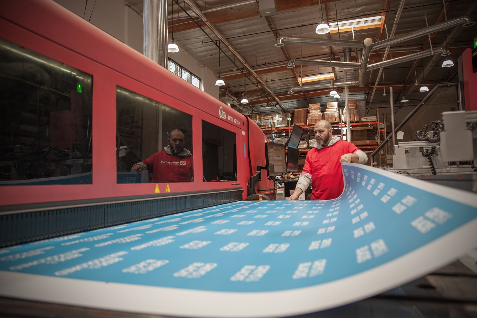 Man in red shirt operating a large printing machine, holding a sheet of printed blue and white labels. Industrial setting.