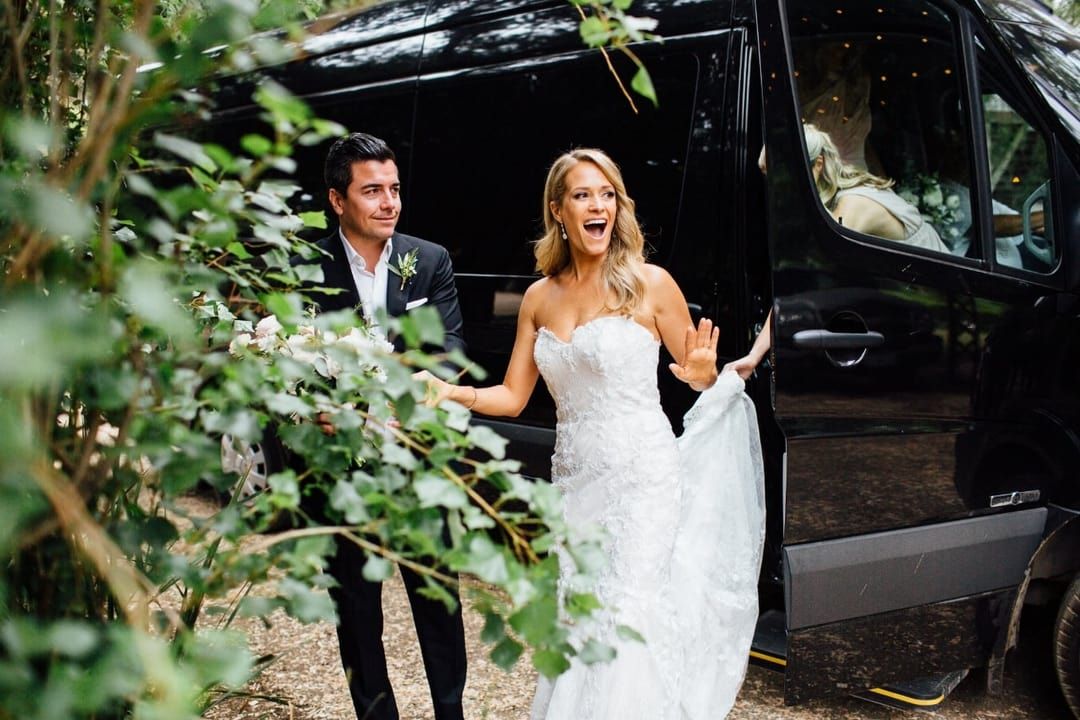Bride and groom exiting black van, bride in strapless white gown, groom in suit, outdoors.