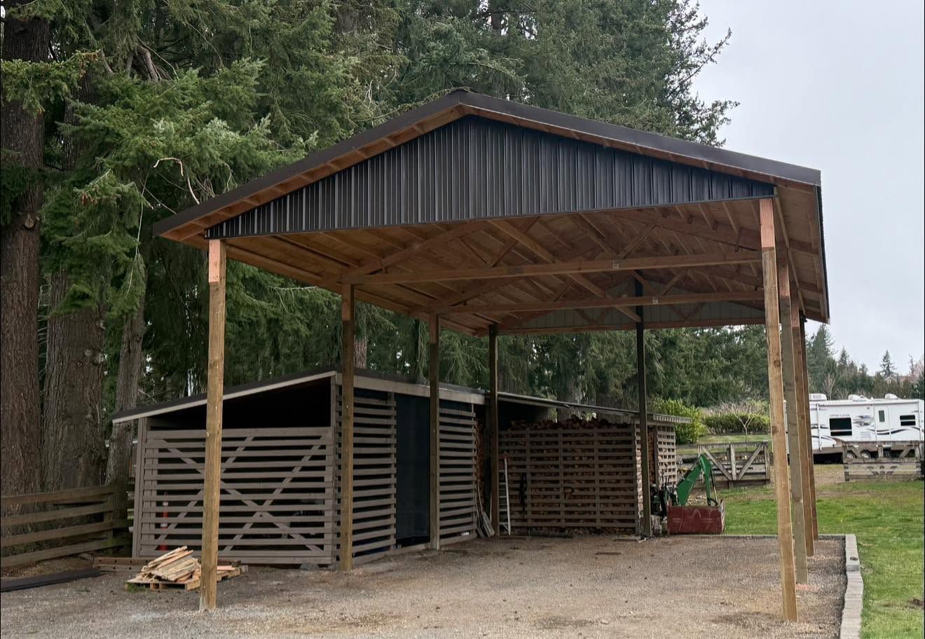 Open-sided wooden livestock shelter with a metal roof beside a gravel area and trees