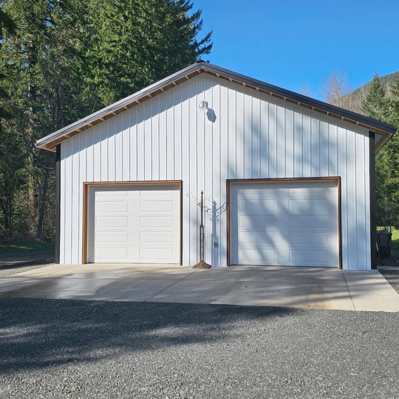 White two-car garage with brown trim and a gravel driveway, set among tall trees under a blue sky