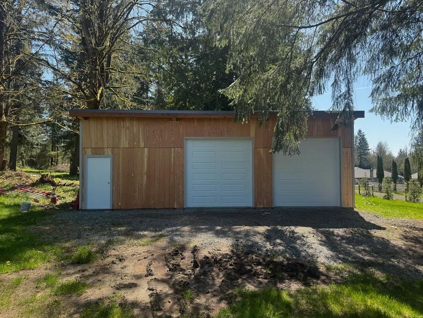 Small wooden shed with two white garage doors and a side door in a wooded yard.