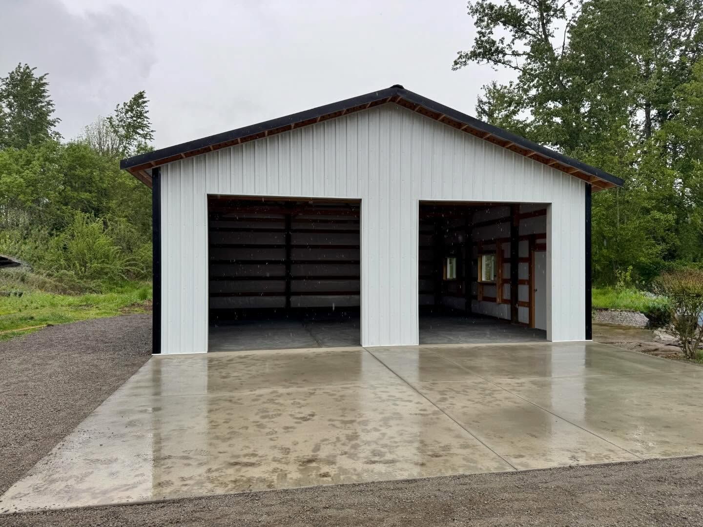 White two-bay garage with open doors on a wet concrete driveway, surrounded by trees.
