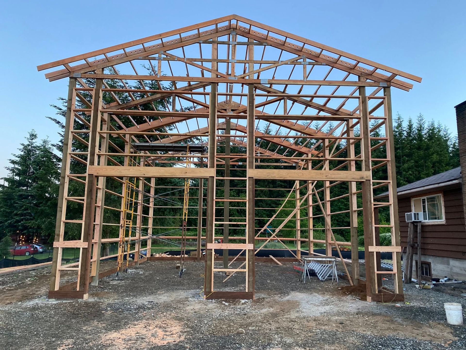 Partially built wooden barn frame on a gravel site, with trees and a house in the background
