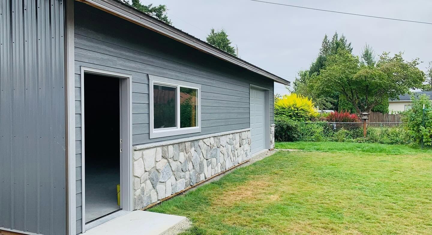 Gray house exterior with open side door, stone lower wall, and a grassy yard with trees and flowers.