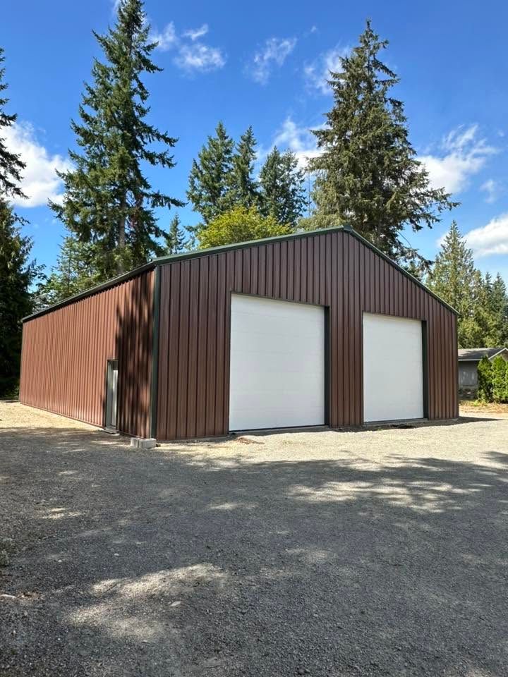 Brown metal garage with two white doors on a gravel lot, surrounded by tall pine trees under a blue sky
