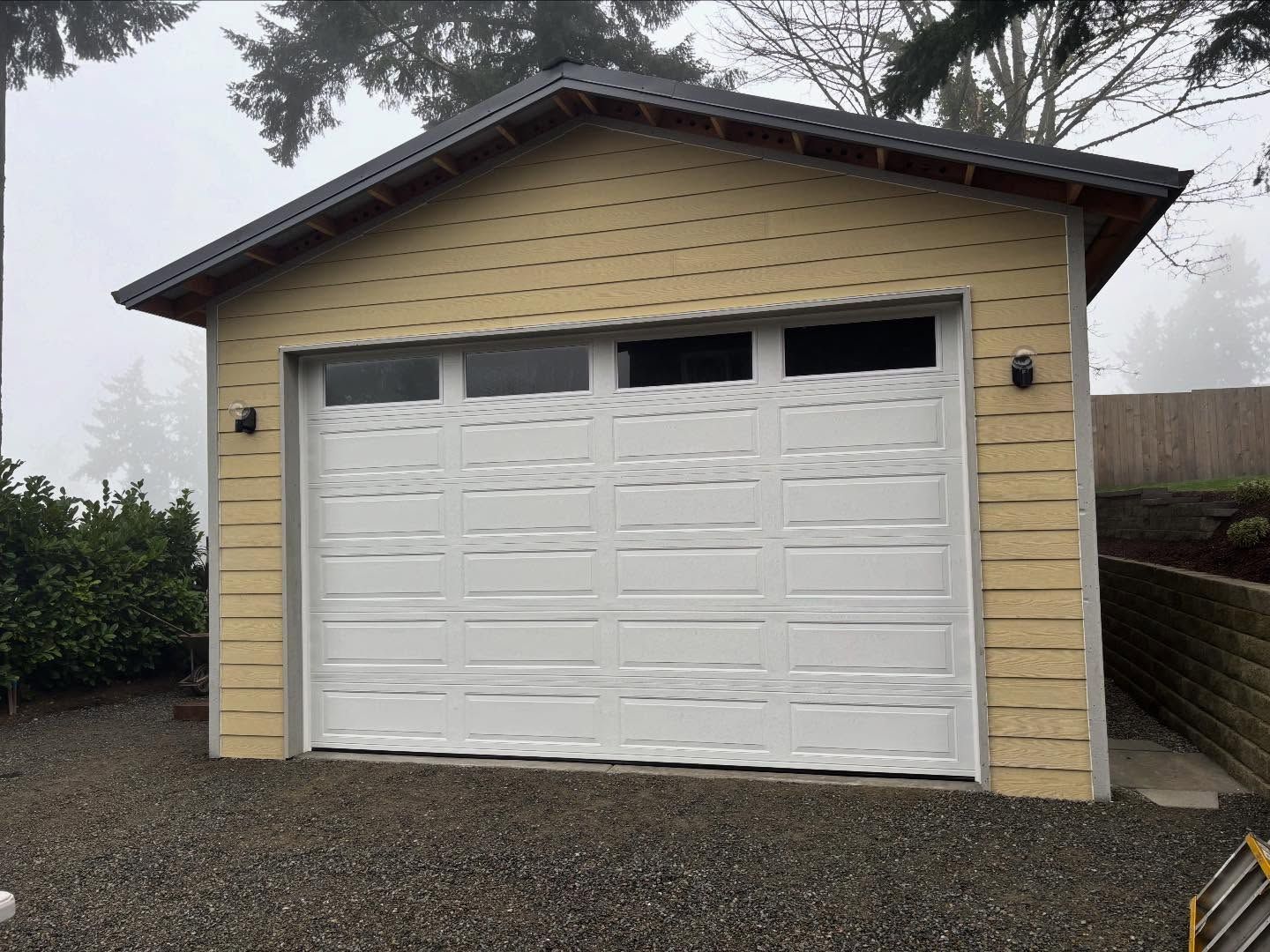 Detached beige garage with a white double door and dark roof on a gravel driveway