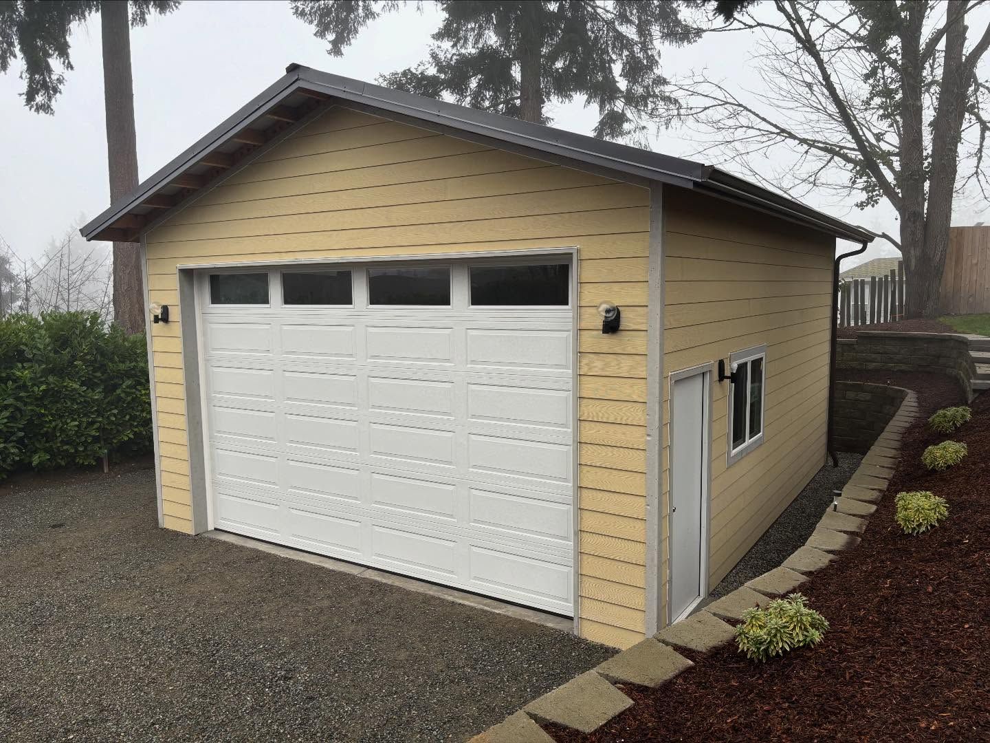 Beige detached garage with white door and side window on a gravel driveway.