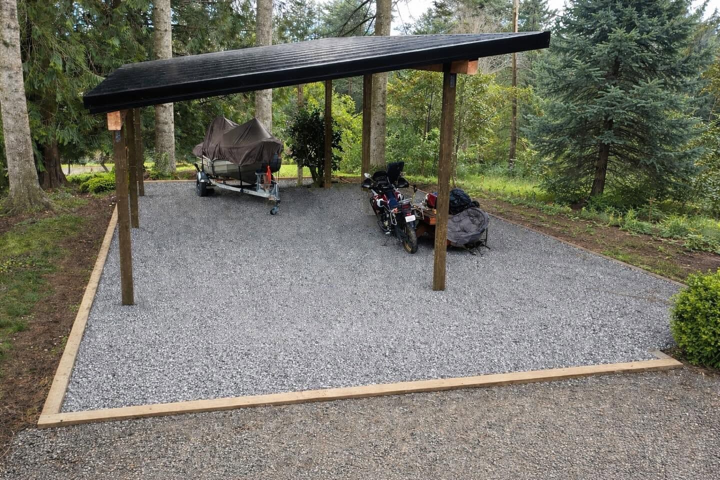 Gravel carport with wooden posts, sheltering a boat and motorcycles in a wooded area.