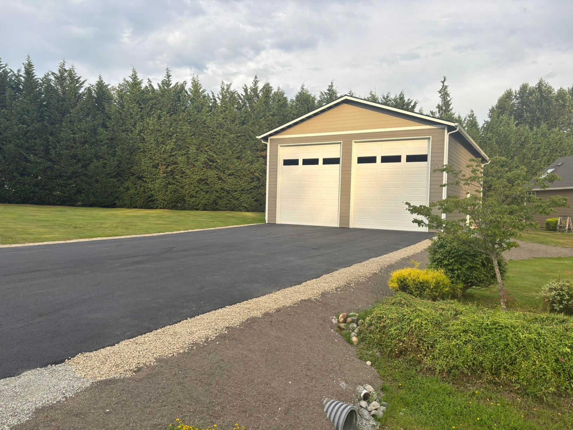 Detached beige garage with two doors beside a paved driveway and gravel landscaping, framed by tall evergreens