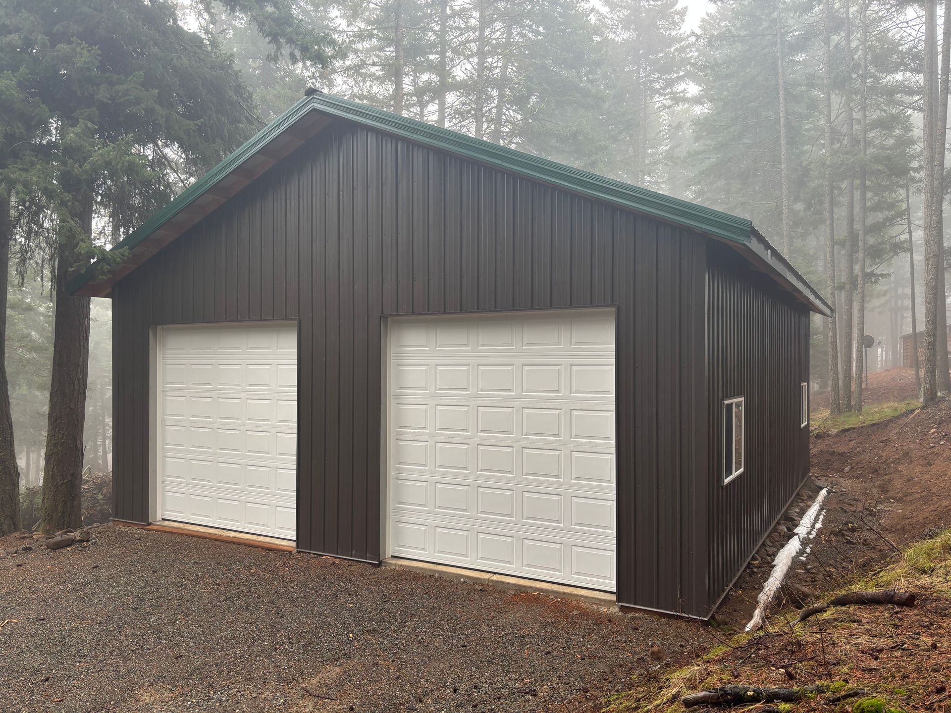 Gray garage with three open bays, parked vehicles, and a gravel driveway surrounded by trees