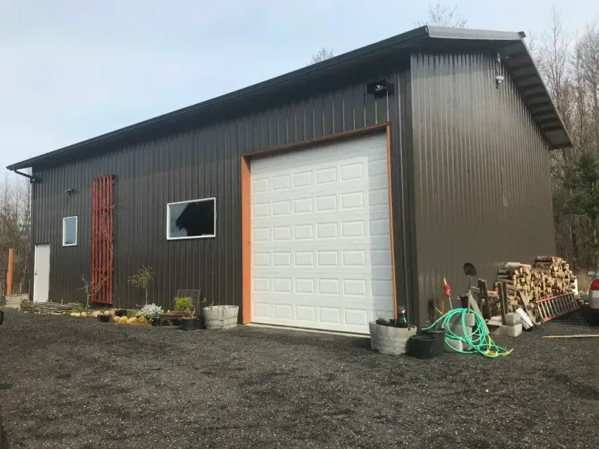 Detached beige double garage beside a black driveway, with gravel landscaping and a tree line in back.