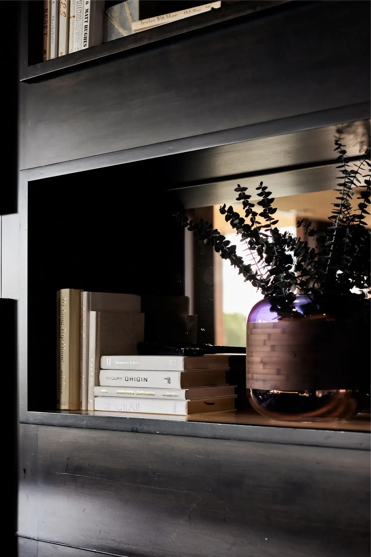 Dark built-in bookshelf with stacked books and a purple vase with greenery.
