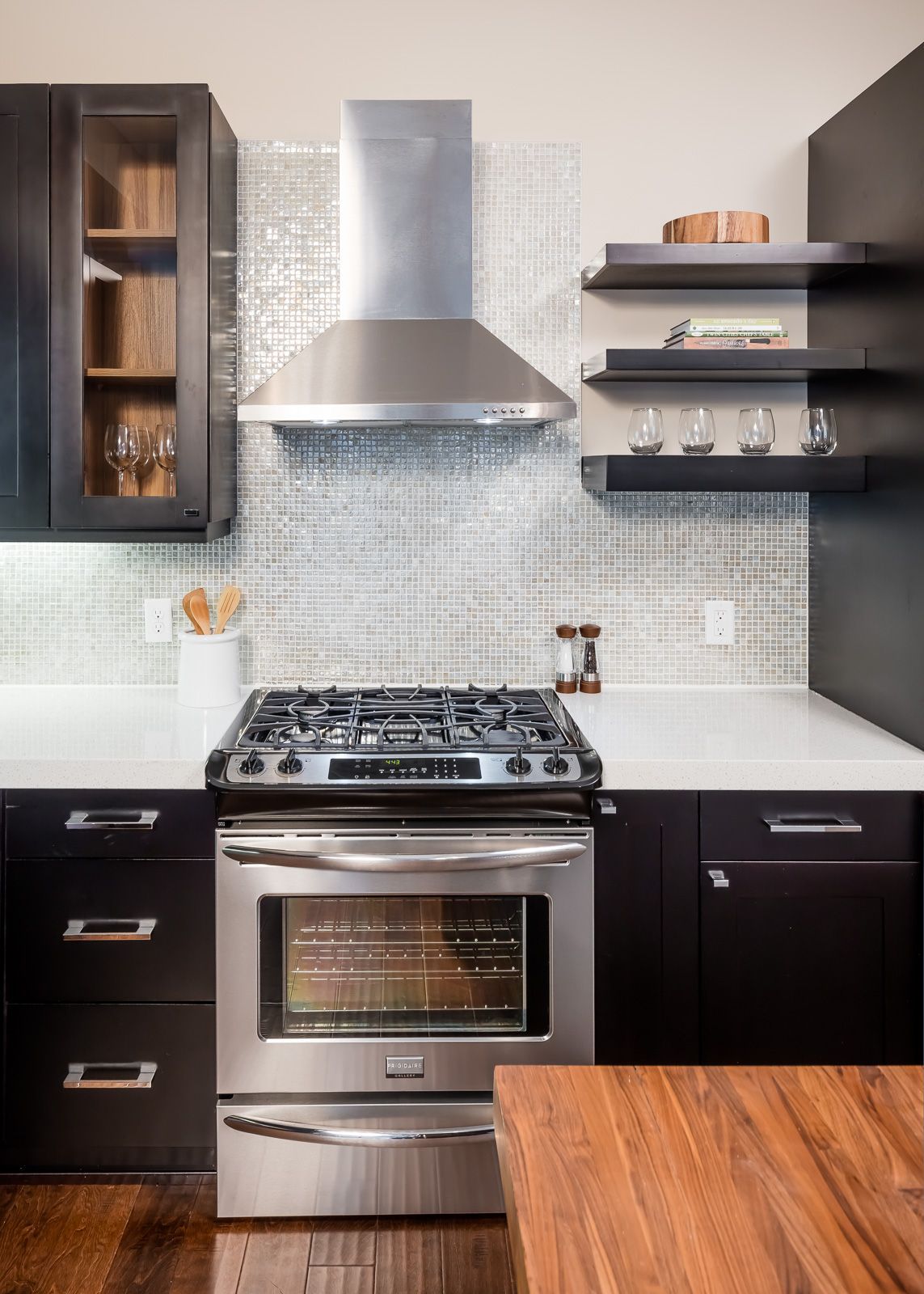 Modern kitchen with stainless steel oven and range hood, black cabinets, and mosaic tile backsplash.