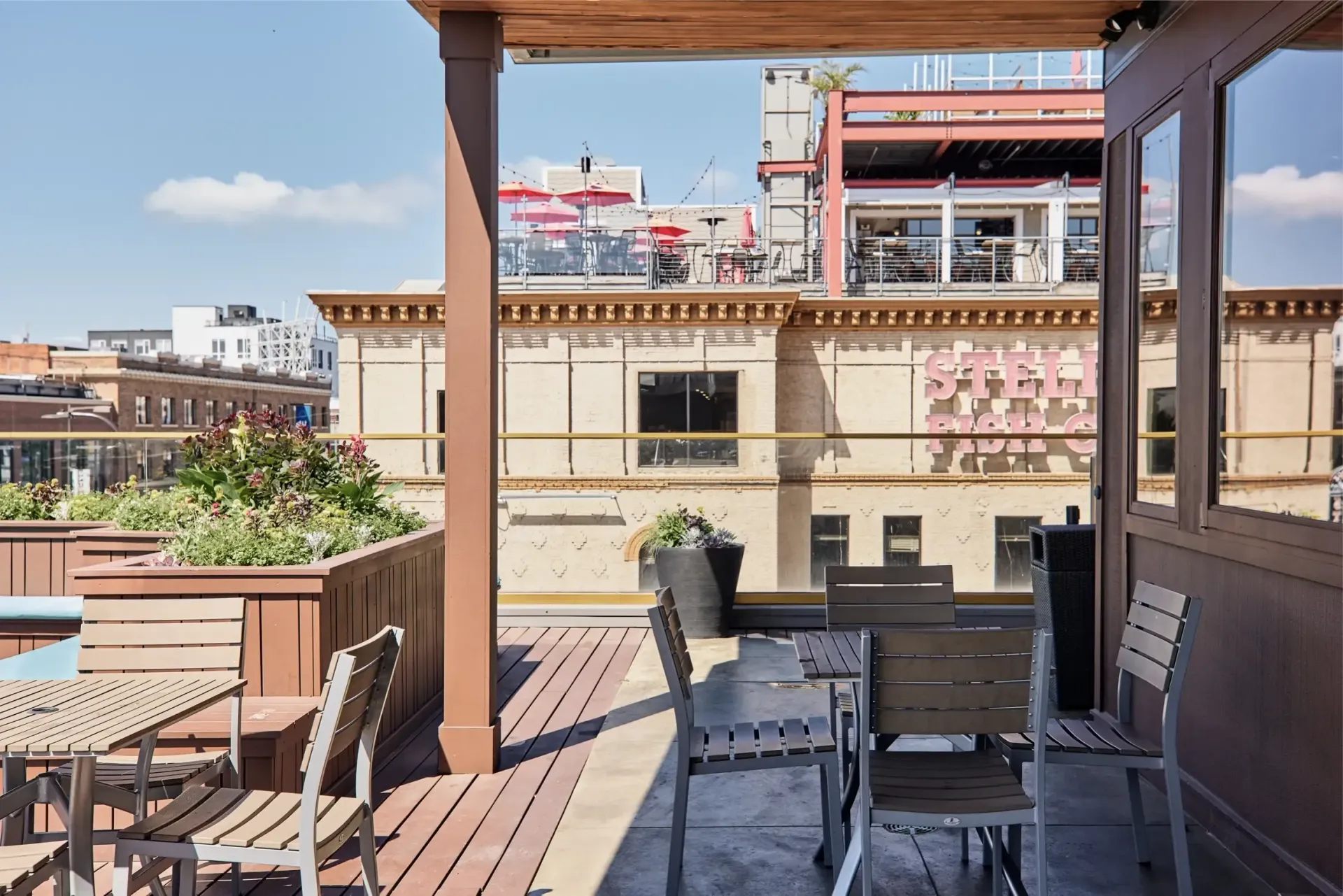 Rooftop communal terrace with tables, chairs, planters, and city buildings in the background.