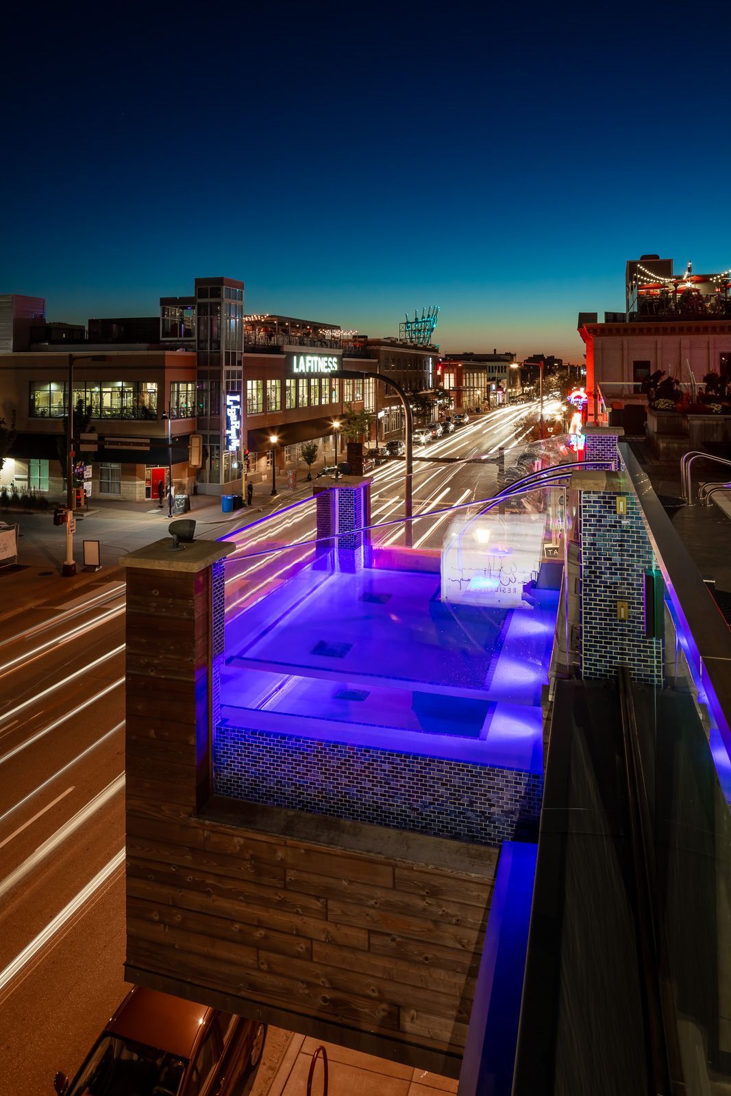 Rooftop pool with blue lighting overlooking a city street at dusk.