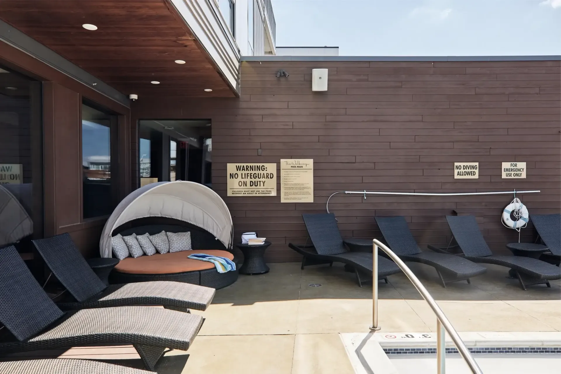 Poolside lounge area with wicker chaise lounges, a curved canopy daybed, and signs on a wood-paneled wall.