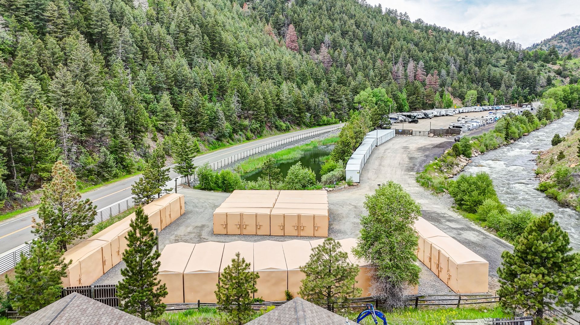 Storage units nestled among trees in a green, mountainous setting along a highway. 