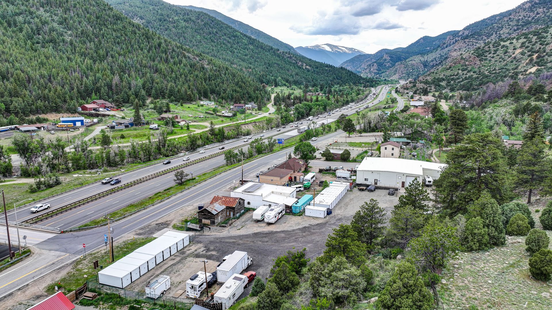 Storage units nestled among trees in a green, mountainous setting along a highway.