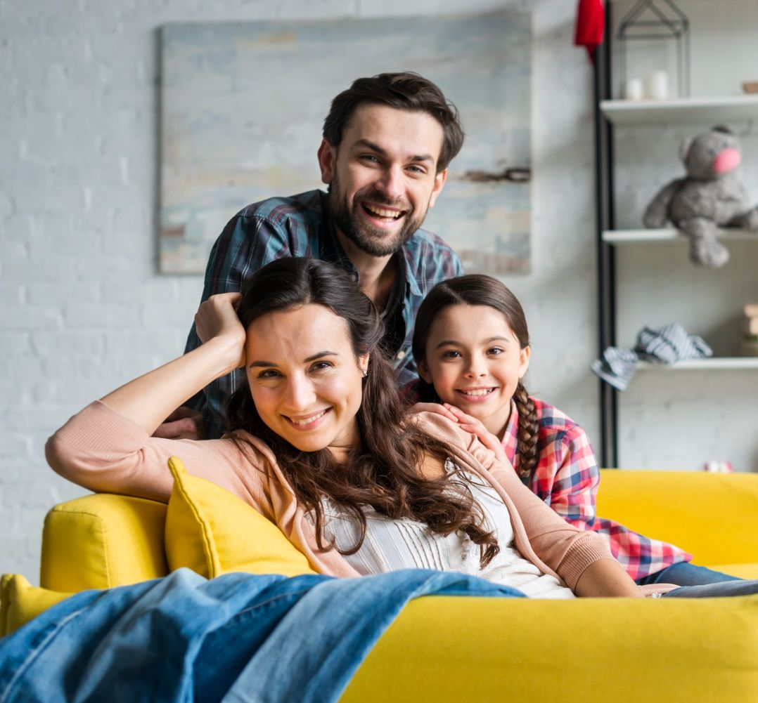 Three smiling people on a yellow couch in a bright living room, with a teddy bear on a shelf behind them