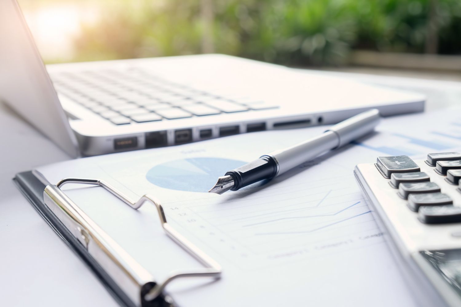 Clipboard, pen, laptop, and calculator on a bright office desk