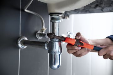 A person uses a bright orange pipe wrench to tighten or loosen a chrome P-trap pipe under a bathroom sink.