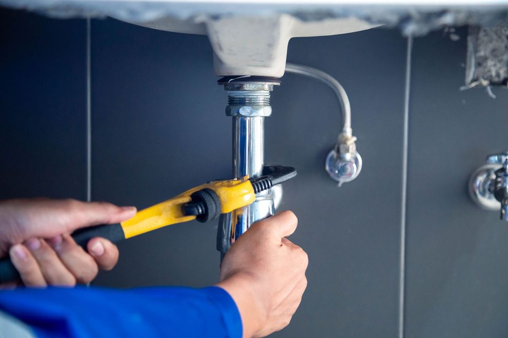 A person in a blue uniform uses a yellow pipe wrench to tighten a chrome drain pipe under a bathroom sink.