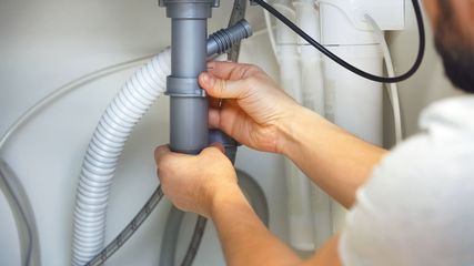 Hands tightening a grey plastic slip-nut joint on plumbing pipes under a sink.