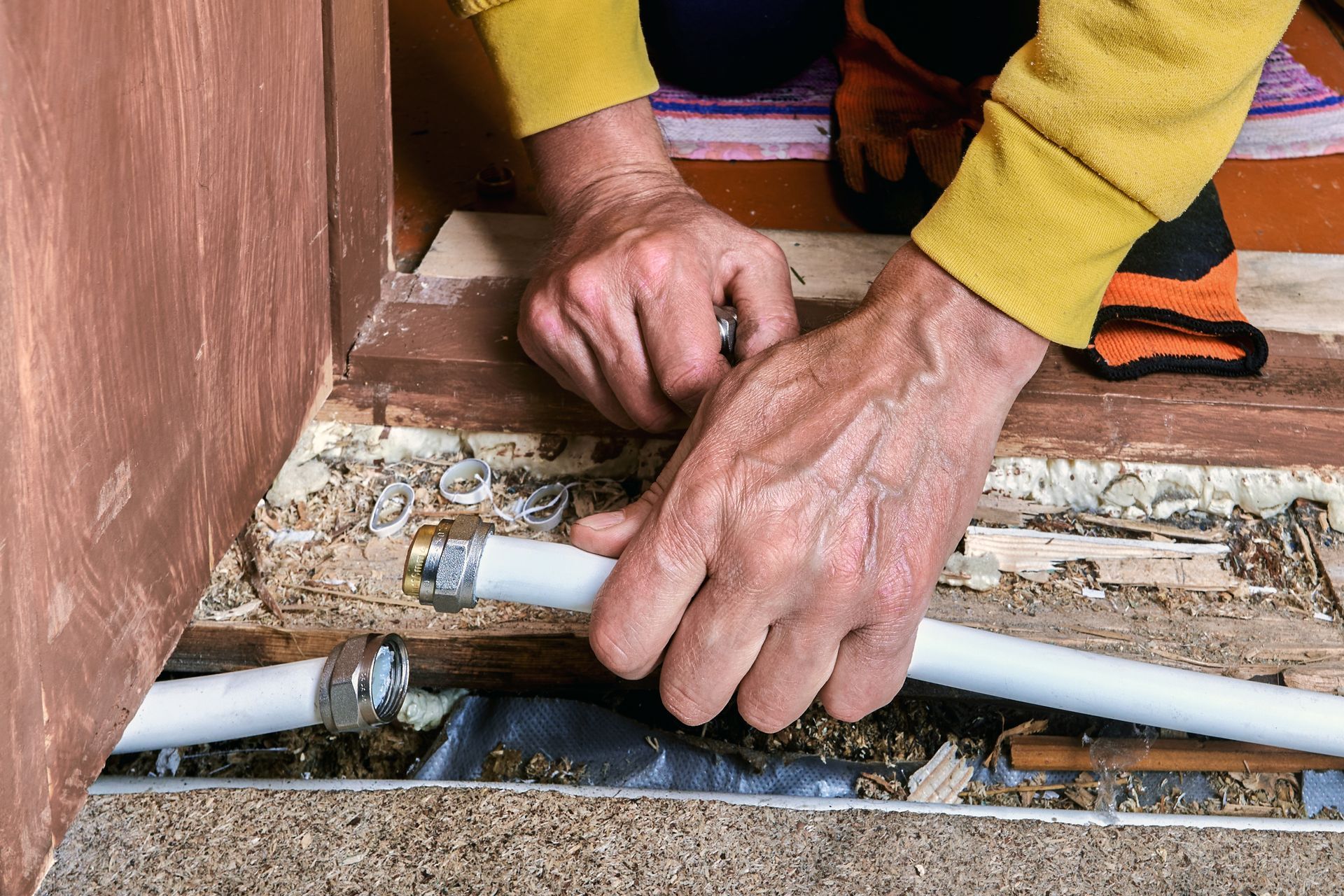 Hands connect two segments of white piping with metal fittings in a recessed floor channel.