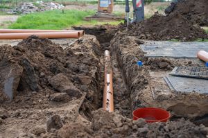 A brown drainage pipe lies inside a narrow excavated trench on a construction site with piles of soil on either side.