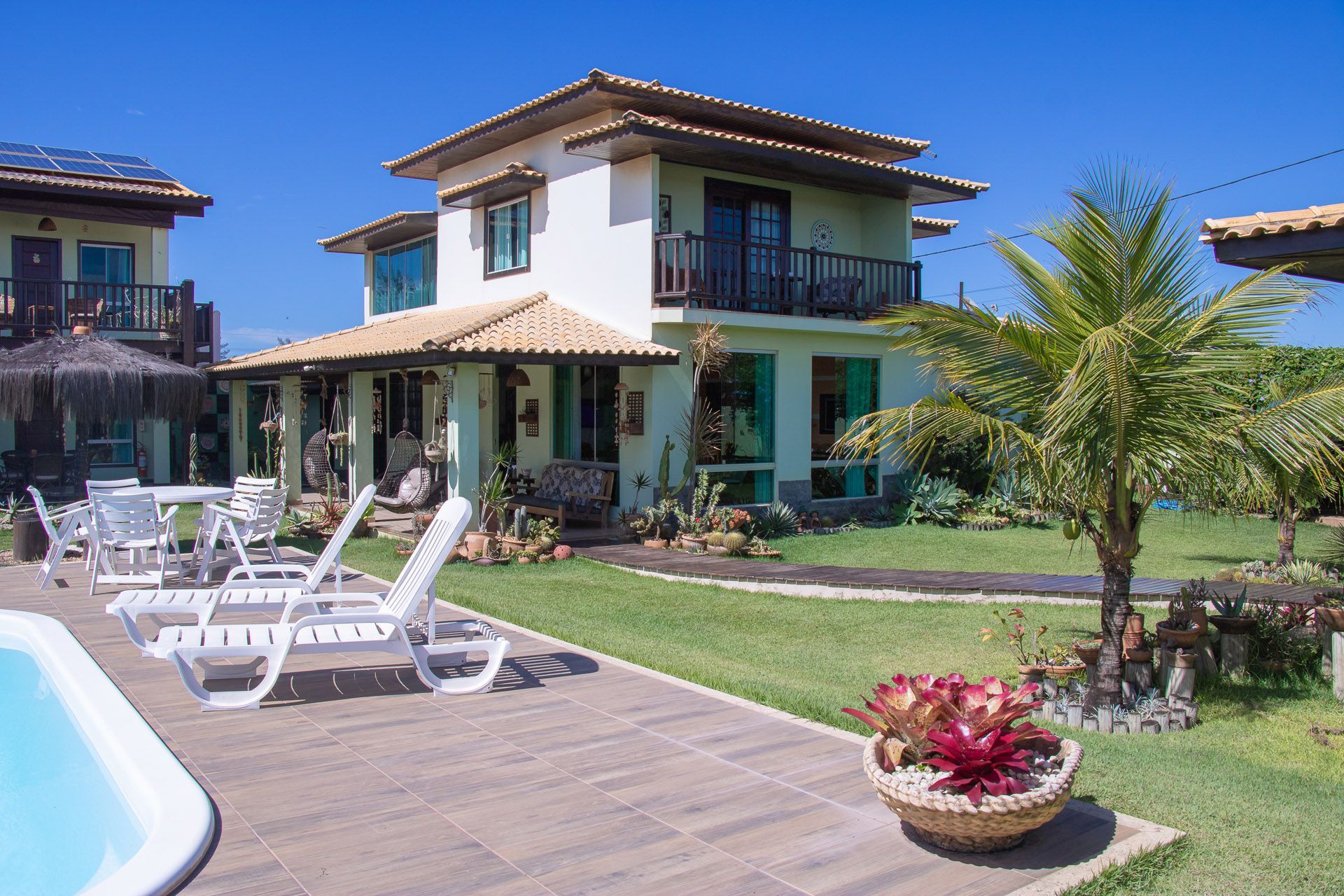Villa luxuosa com piscina, espreguiçadeiras brancas e paisagismo tropical em um dia ensolarado.