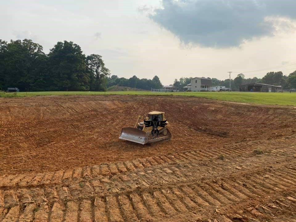 Bulldozer grading a large dirt field under a cloudy sky, with trees and buildings in the distance
