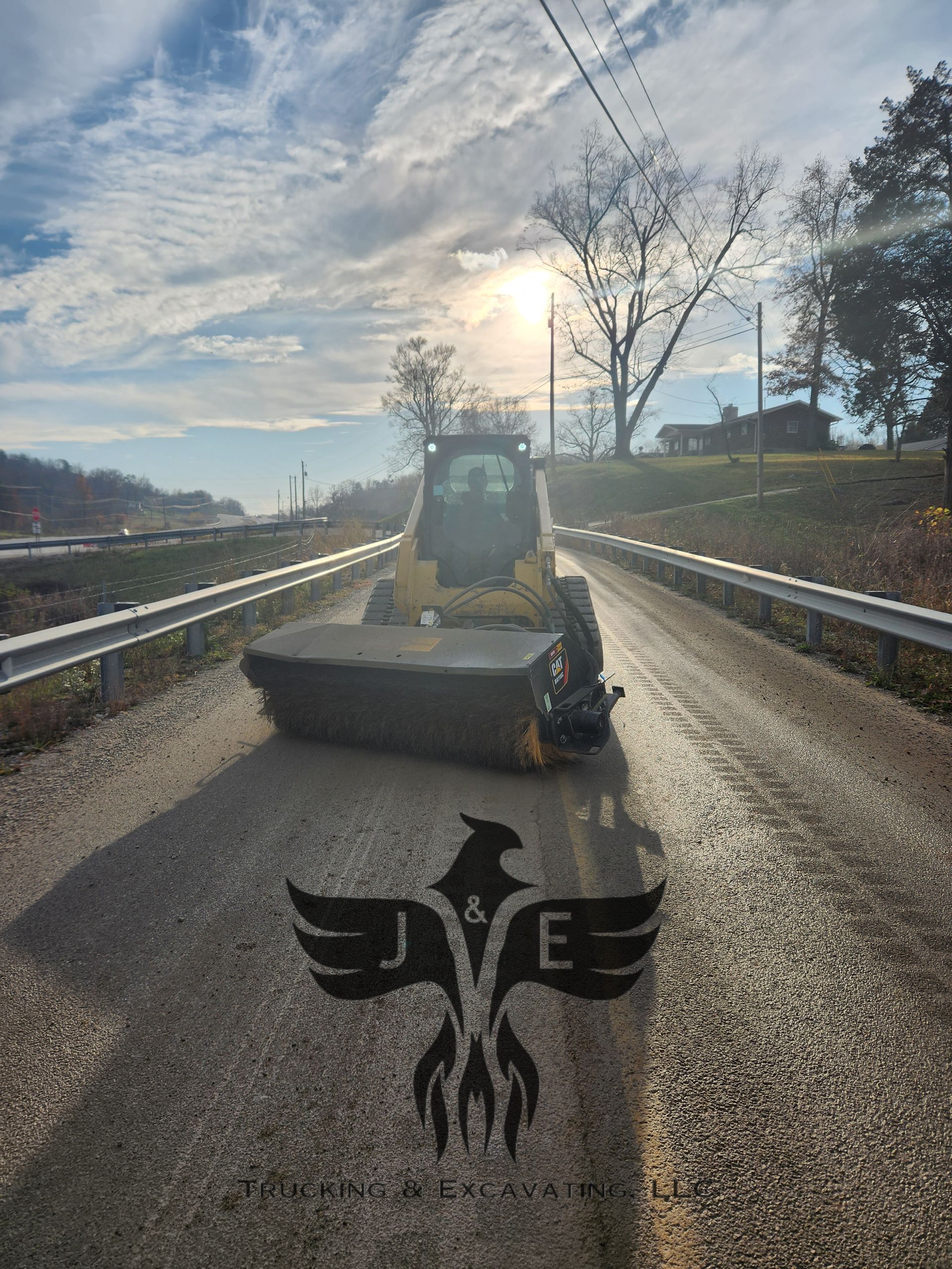 Road roller on a wet road at sunrise, with a winged emblem in the foreground.