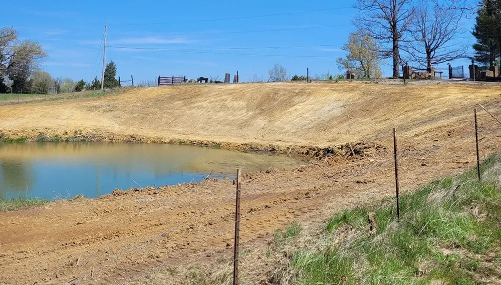 Dry grassy hill beside a small pond under a clear blue sky, with fence posts in the foreground.