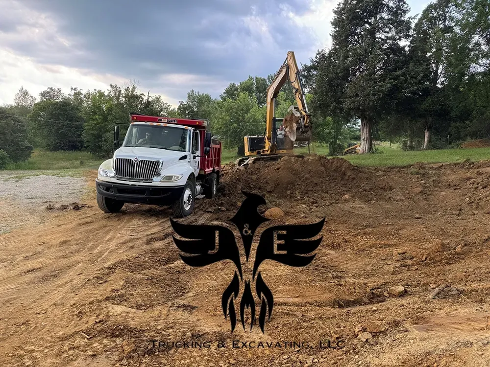 Tow truck and excavator at a muddy roadside worksite, with a black winged emblem in the foreground