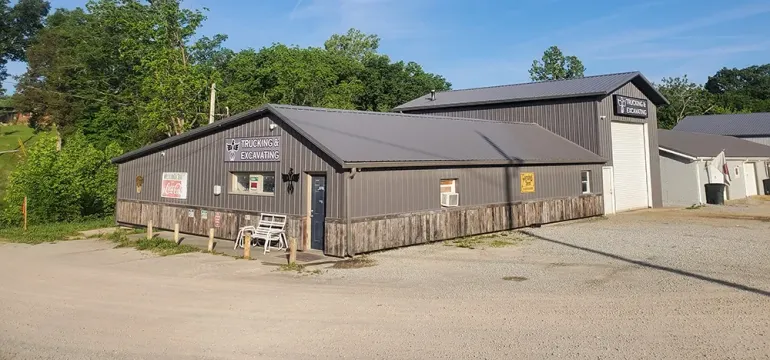 Brown wooden building with a gray roof on a gravel lot beside trees and neighboring structures