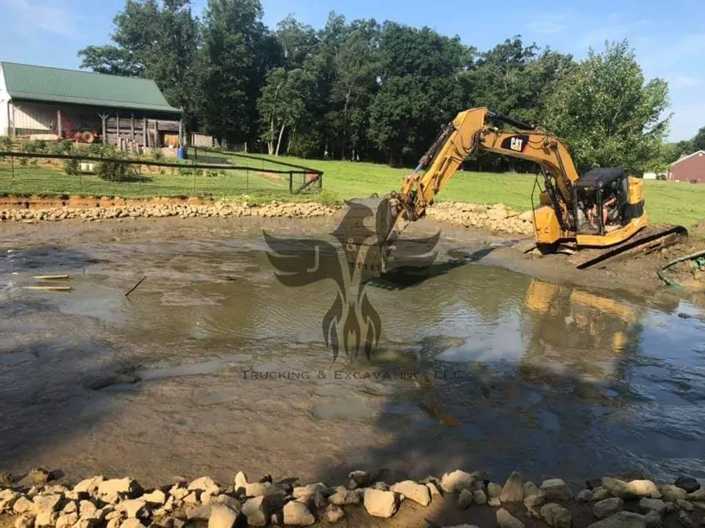 Excavator digging in a muddy pond beside a house and wooded shoreline
