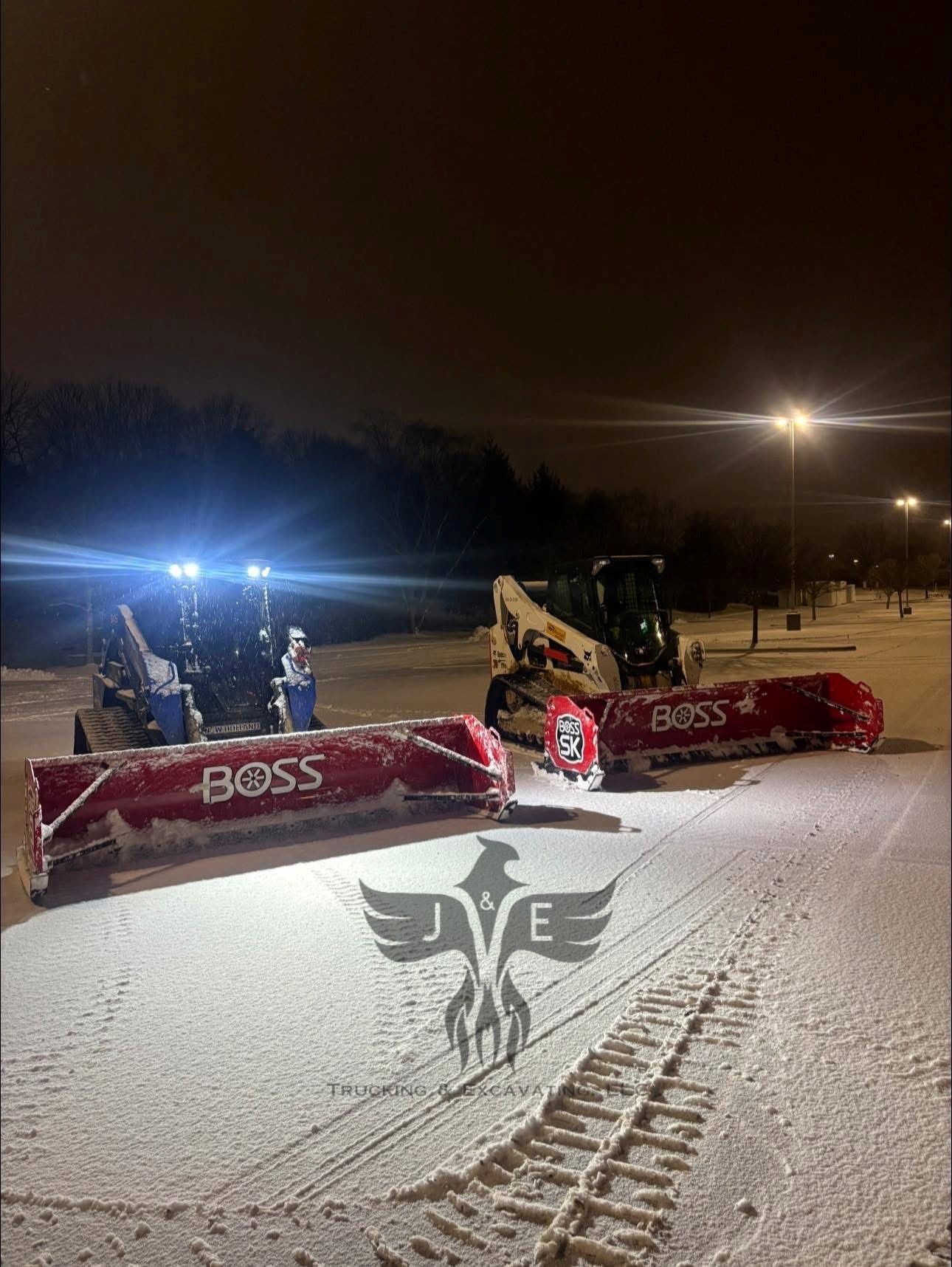 Two snowplows clearing a snowy road at night under bright lights