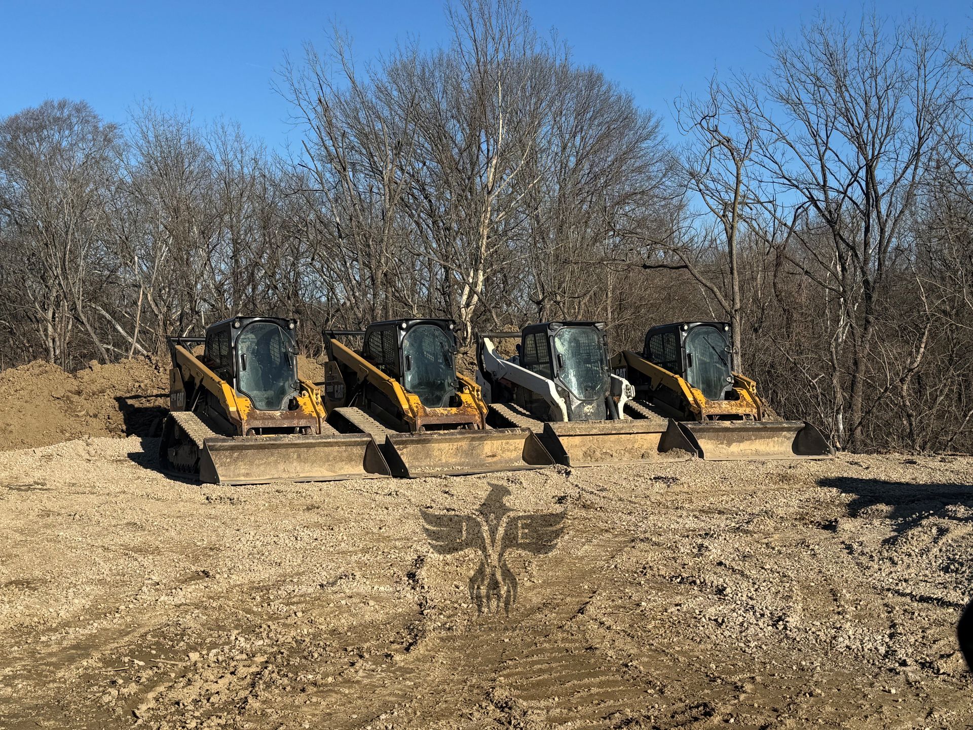 Four construction loaders parked on a dirt lot beside leafless trees on a clear day