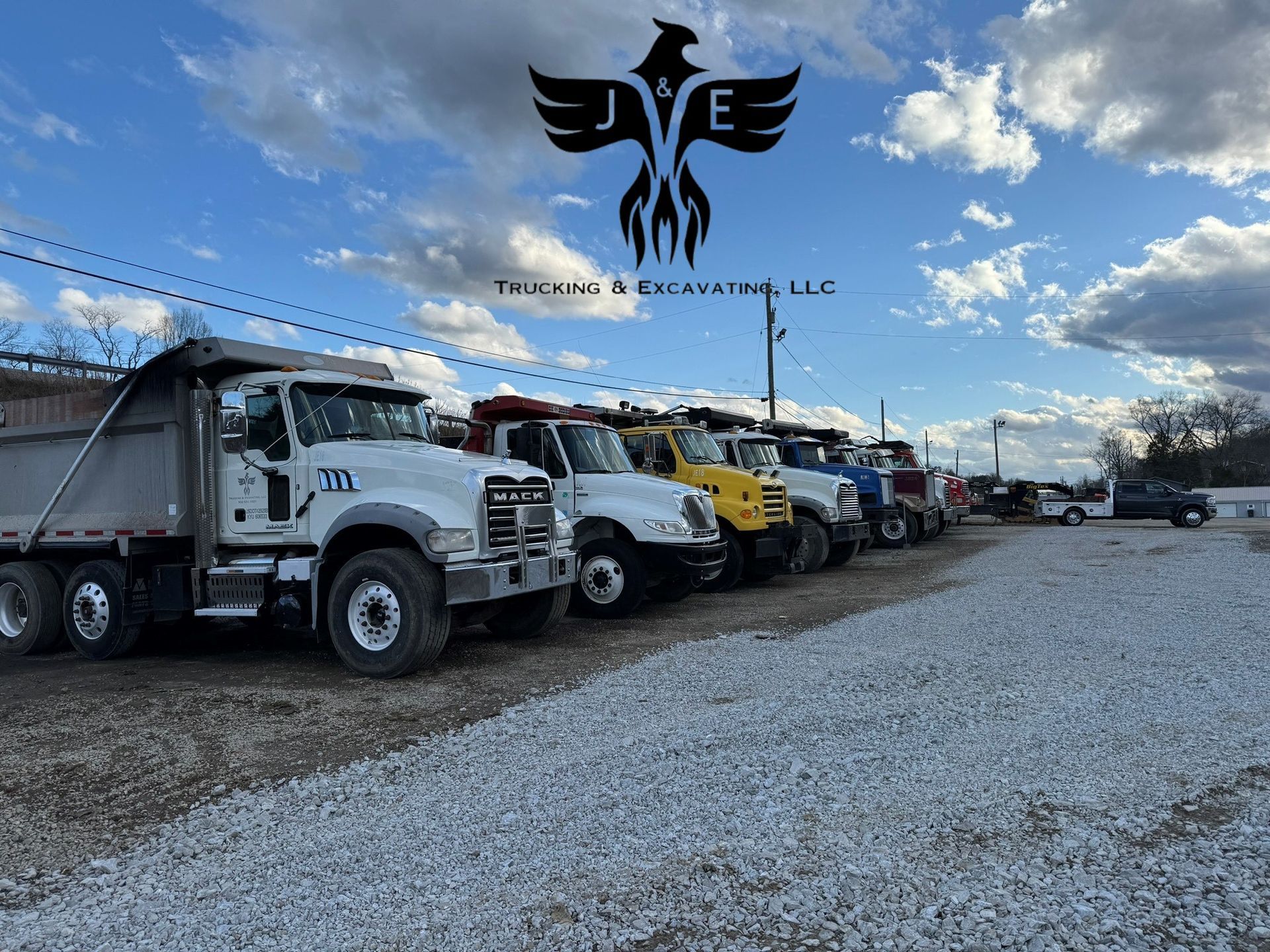 Row of parked dump trucks and utility vehicles on a gravel lot under a cloudy blue sky