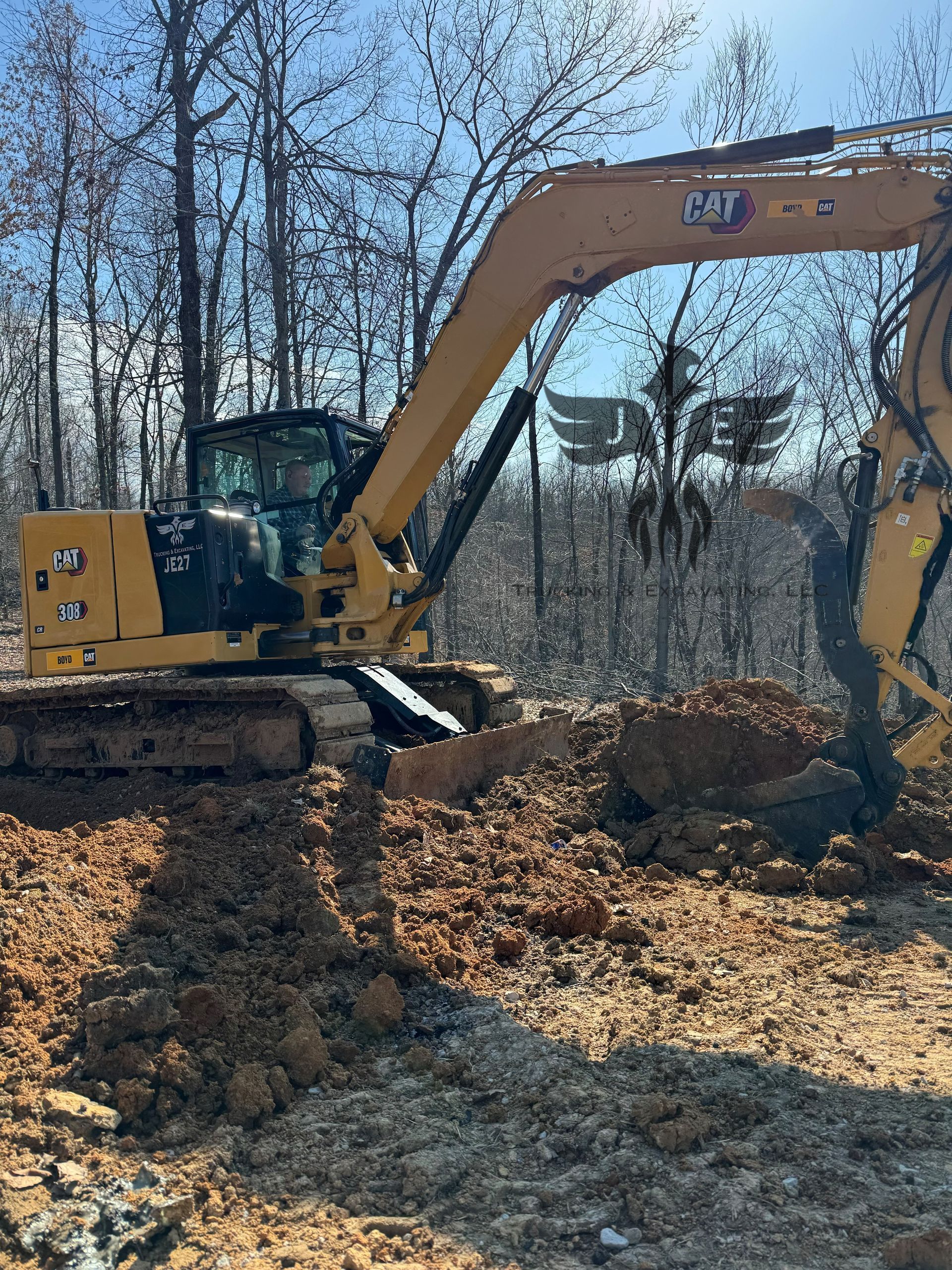 Yellow excavator clearing leaves in a wooded area beside a pile of logs.