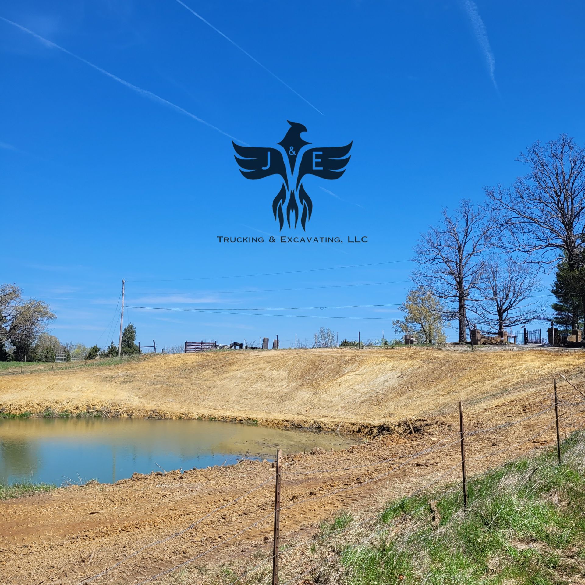 Blue sky over a dry grassy hill beside a pond, with a black winged logo in the sky