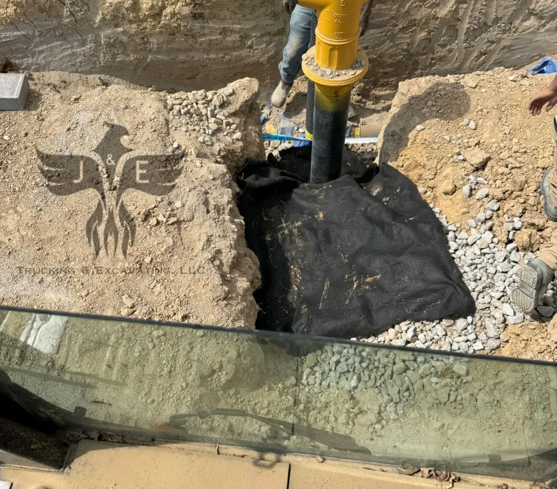 Worker in yellow safety gear standing in a trench beside a black pipe and excavated dirt
