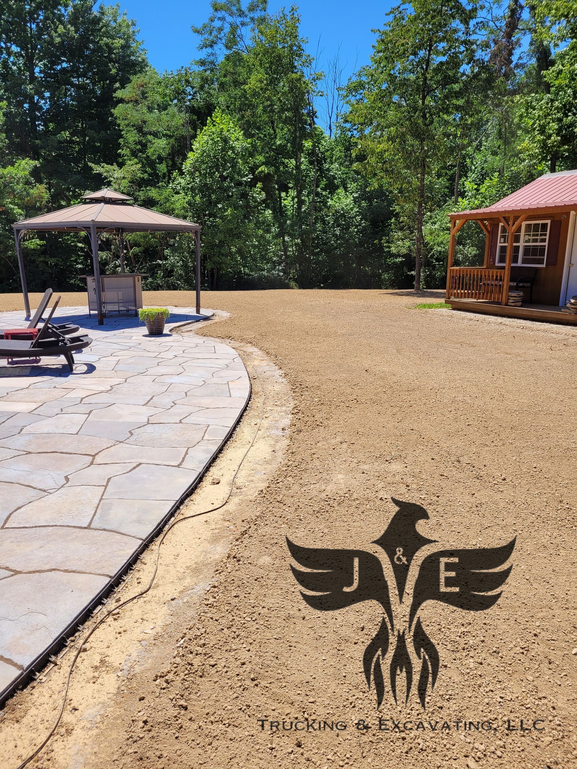 Backyard patio with stone path, umbrella, gazebo, trees, and woodchip ground under a blue sky