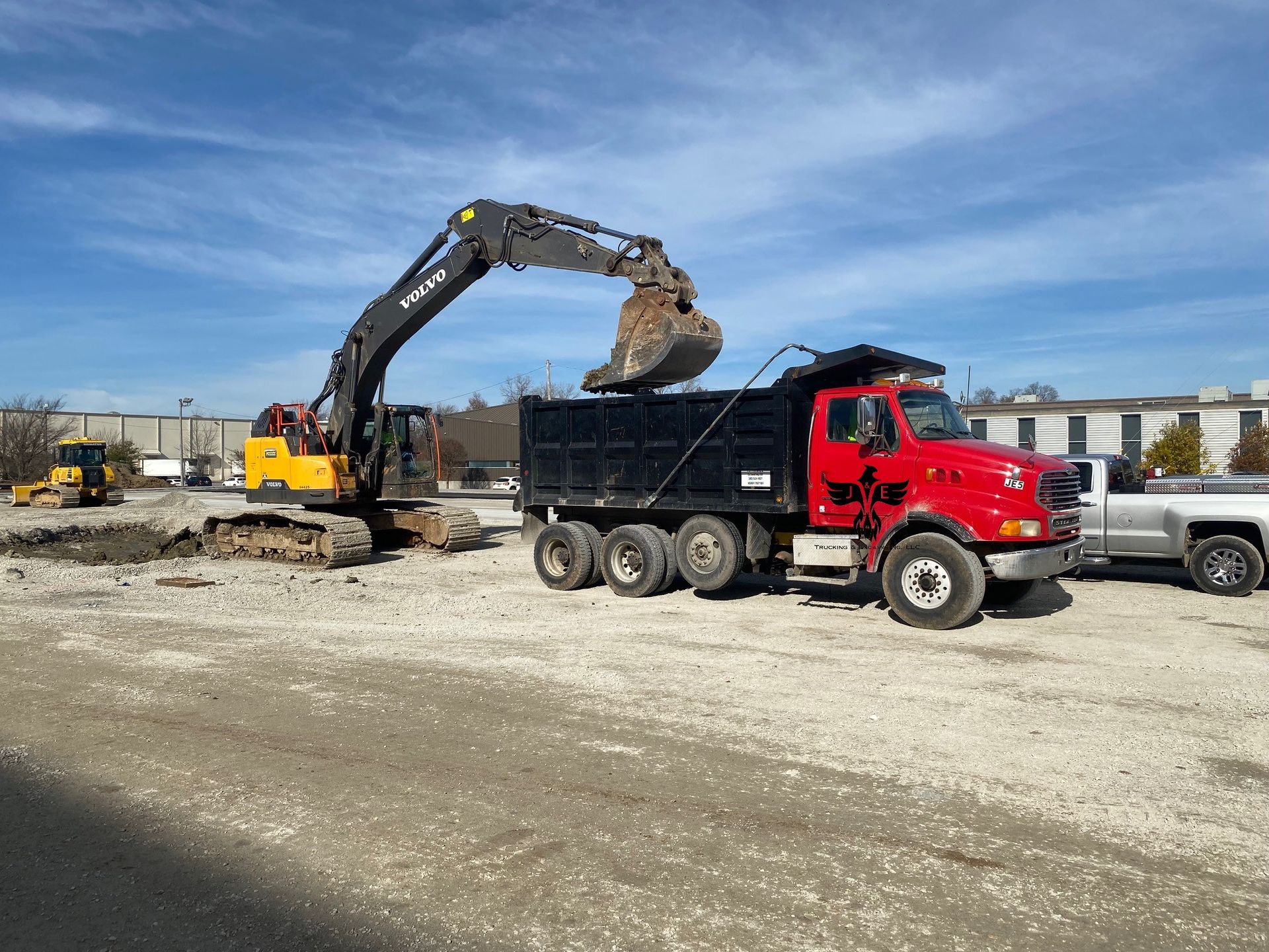 Excavator loading dirt into a black dump truck beside a red truck at a construction site.