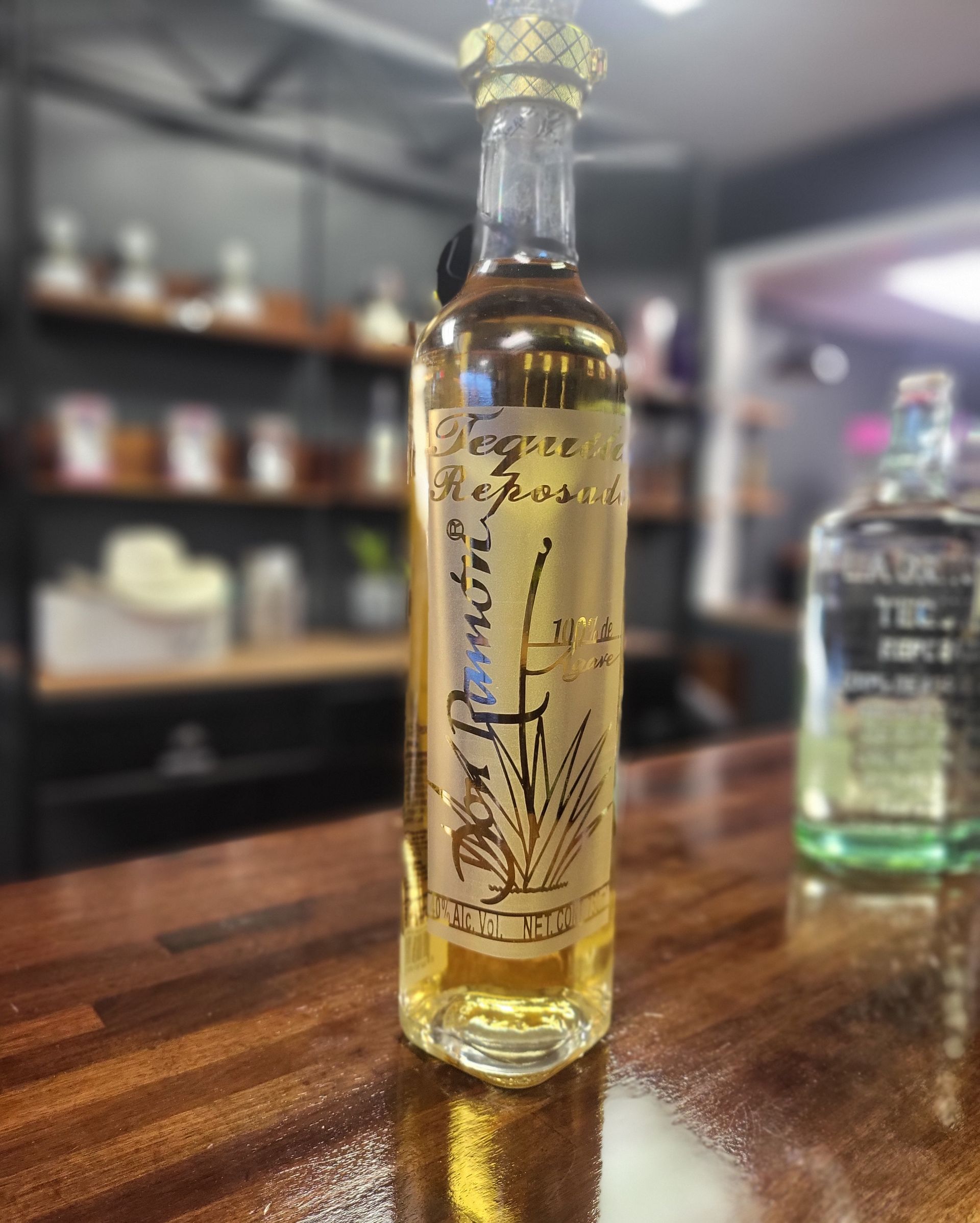 Tall amber bottle with a gold cap on a wooden counter in a bar, with shelves blurred in the background
